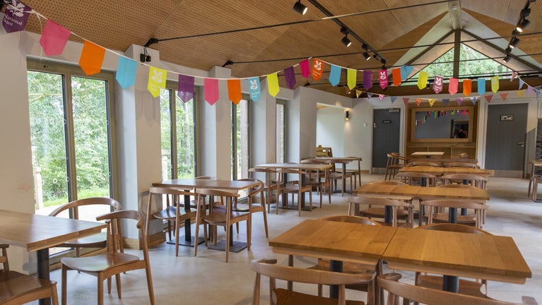 Tables and chairs with windows on the left inside the cafe. There are two doors at the rear of the room. One of them is the accessible toilet. There is bunting hanging from the ceiling.